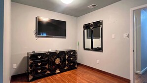 A room with a wooden floor, a flat-screen TV mounted on the wall, a large mirror, and a black entertainment unit with drawers.