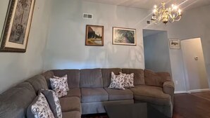 A living room with a sectional sofa, a glass coffee table, and a chandelier. There are three framed pictures on the wall.