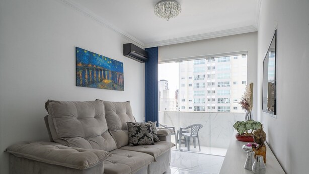 Ein modernes Wohnzimmer mit einem beigen Sofa, einem Gemälde, einem Blumentopf und Blick auf Gebäude durch ein großes Fenster.