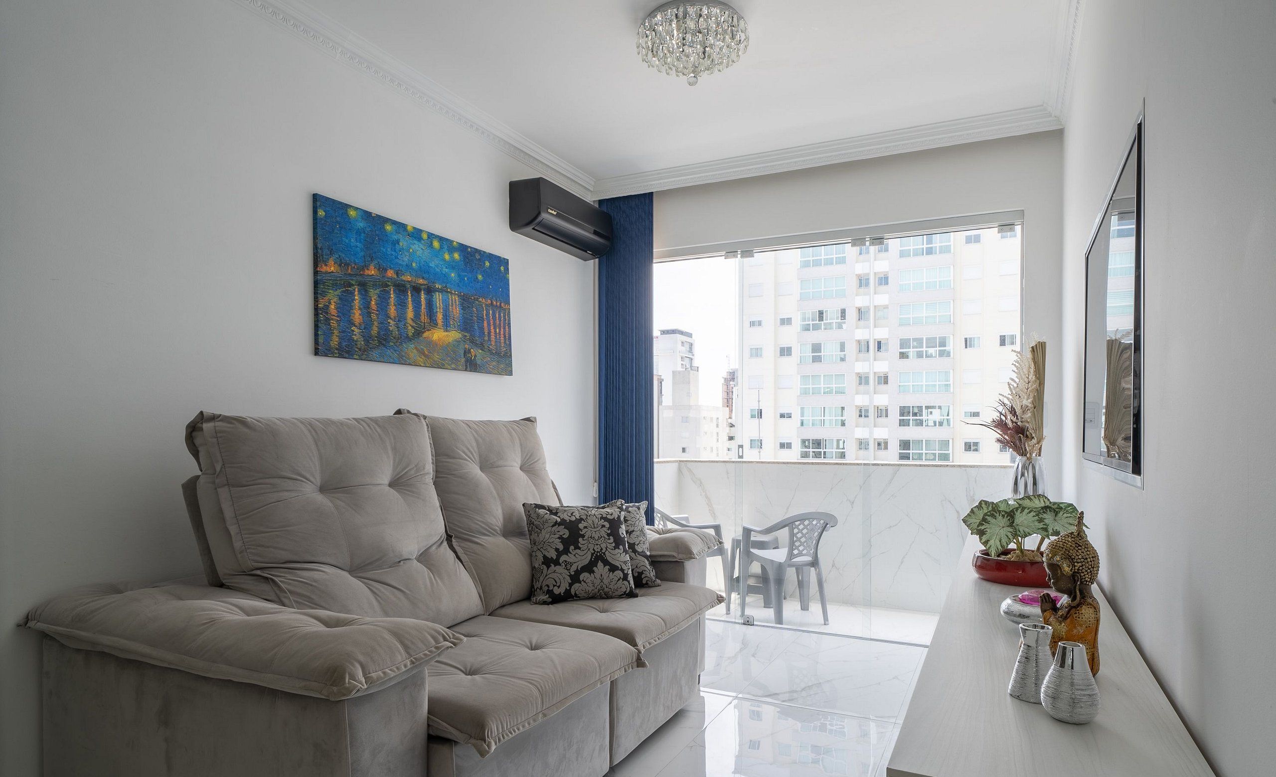 A modern living room with a beige sofa, a painting, a potted plant, and a view of buildings through a large window.