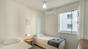 Una stanza piccola e pulita con due letti, un comodino con una lampada, una finestra vista edifici e una ventola a soffitto.