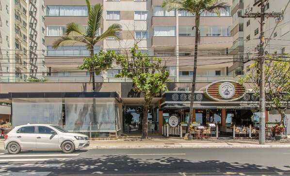 Une vue de rue avec une voiture blanche garée sur le bord, un immeuble abritant un restaurant appelé « PARRILLA ARGENTINA » avec une terrasse, et un grand immeuble d’habitation en arrière-plan.