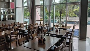 Un restaurante espacioso con mesas y sillas de madera, ventanas amplias con cortinas y vistas a palmeras y una piscina al exterior.