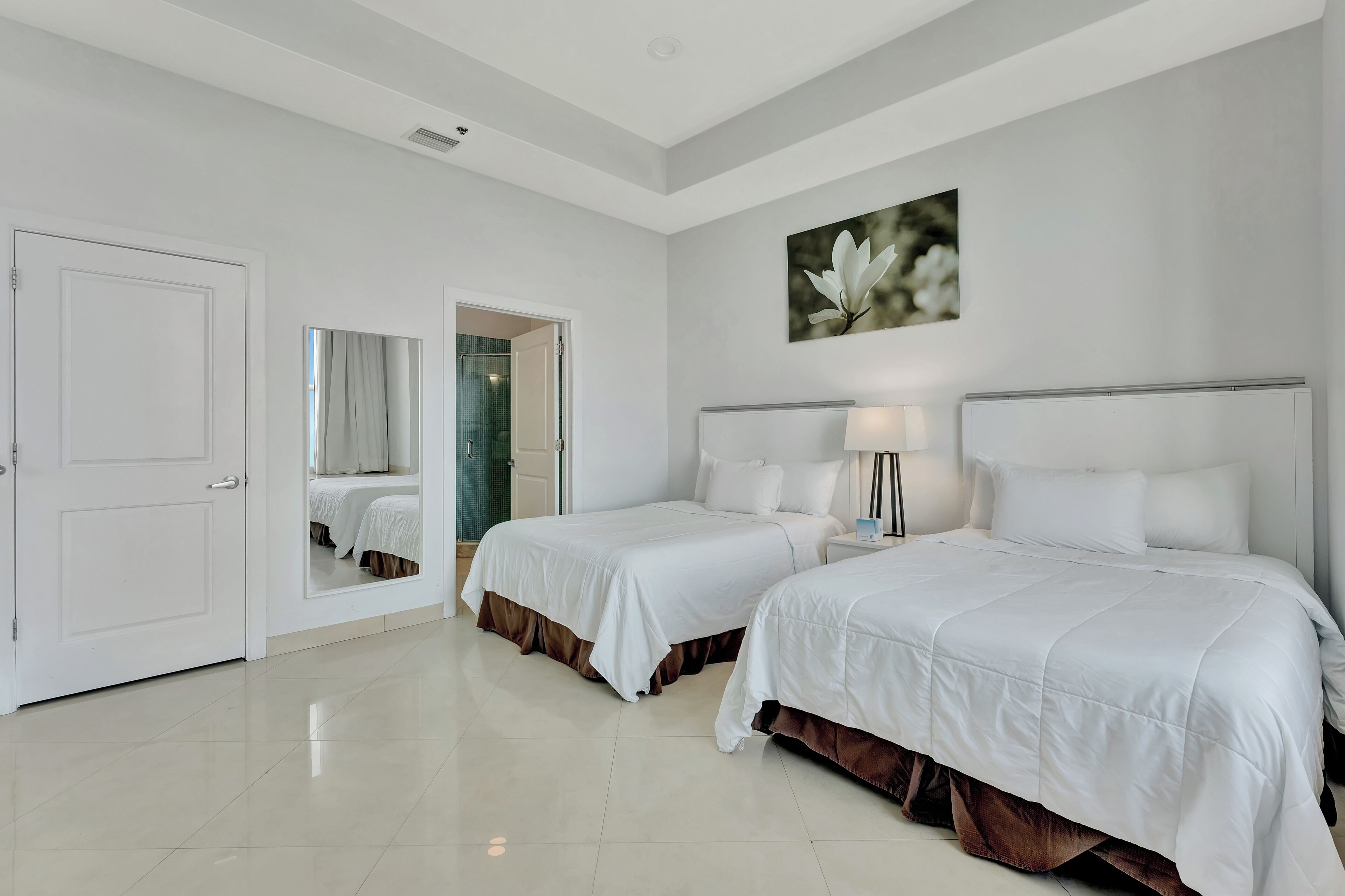 Una habitación de hotel con dos camas, un espejo grande, un cuadro y una puerta que da a un baño.