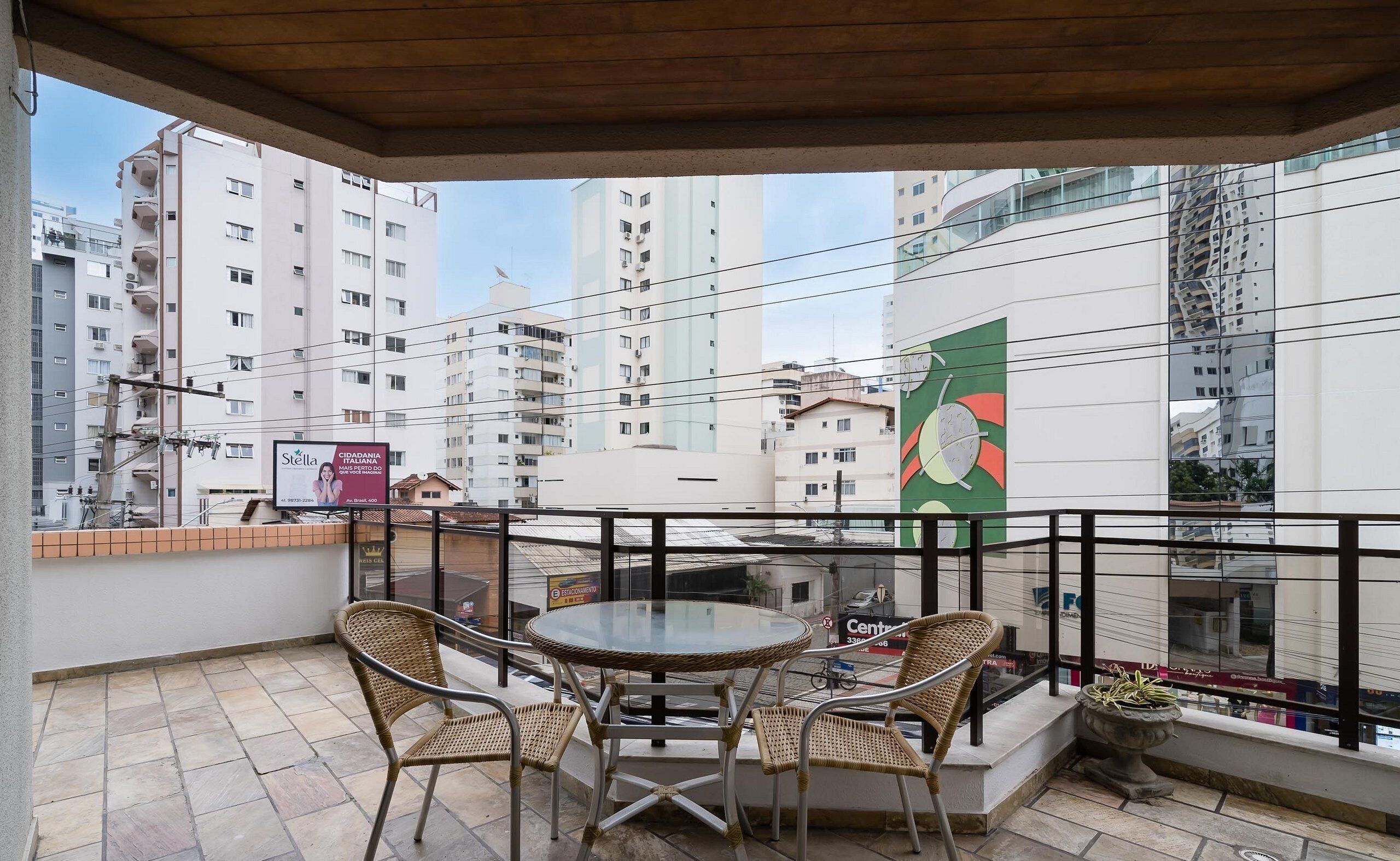 Un balcón con sillas de mimbre y una mesa de vidrio, con vistas a un paisaje urbano con edificios modernos y un gran cartel publicitario.