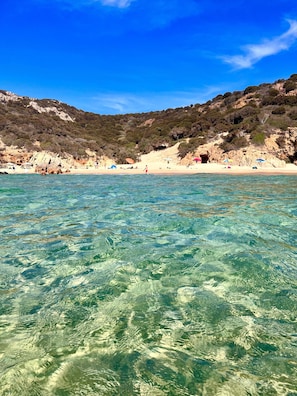 A beach with clear water, sandy shore, and rocky hills in the background.