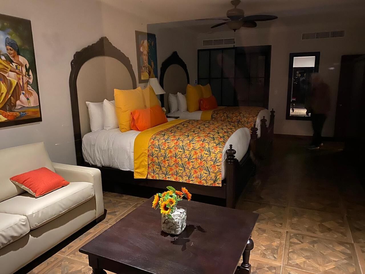 A hotel room with two beds, a sofa, a coffee table with a vase of flowers, and a ceiling fan.