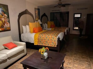 Ein Hotelzimmer mit zwei Betten, einem Schlafsofa, einem Couchtisch mit Blumenvase und einem Deckenventilator.