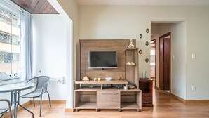 Un salon moderne avec un meuble de rangement en bois pour l’équipement audiovisuel, un téléviseur à écran plat et un coin repas comprenant une table en verre et des chaises en métal.
