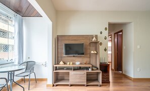 Uma sala de estar moderna com um móvel de entretenimento de madeira, uma TV de tela plana e um espaço de jantar com mesa de vidro e cadeiras de metal.