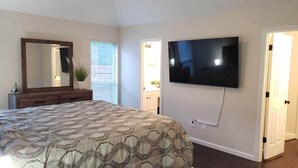 A bedroom with a large bed, a dresser, a mirror, a TV mounted on the wall, and a door leading to another room.