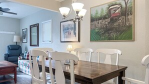 A dining area with a wooden table and white chairs, framed artwork on the walls, and a ceiling fan.