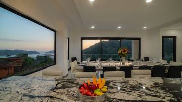 A modern dining area with a marble table, a fruit bowl, and a view of a mountainous landscape through large windows.