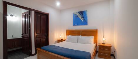 A bedroom with a wooden bed, a nightstand with a lamp, a ceiling fan, and a painting on the wall.