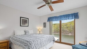 A bedroom with a bed, a nightstand, a lamp, a ceiling fan, a window with a view of a lake, and a blue upholstered chair.
