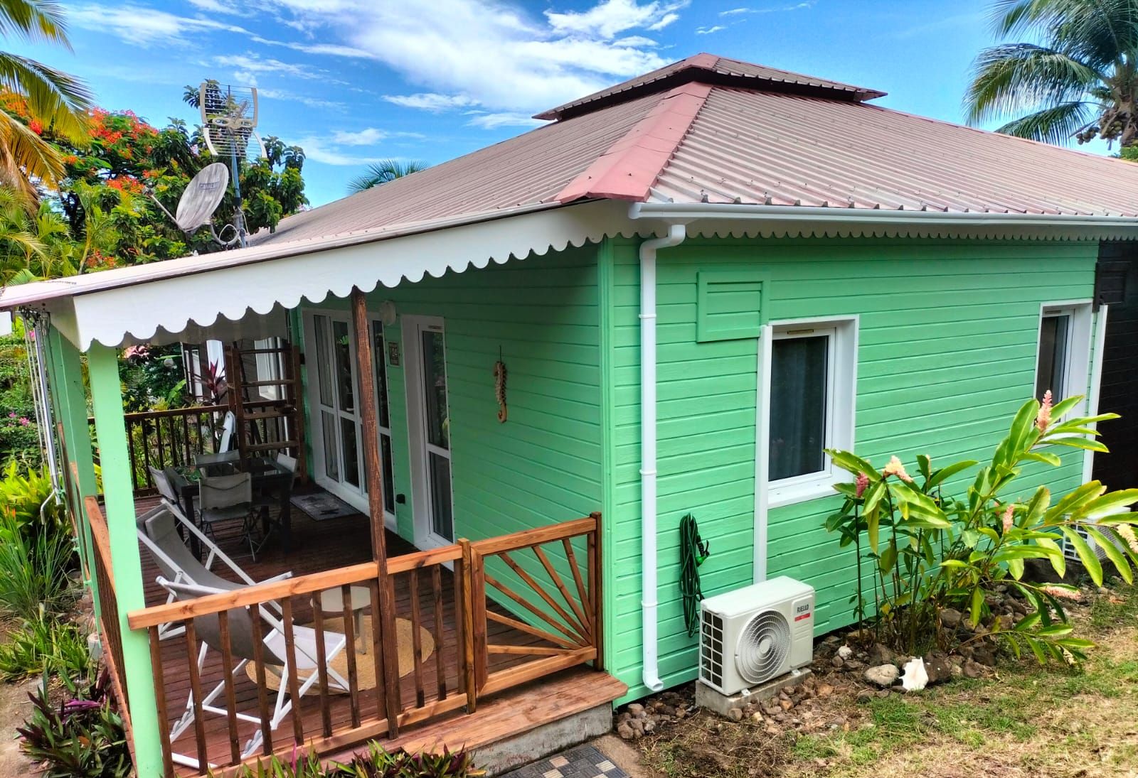 A small, single-story house with a green exterior and a red roof. It features a porch with a wooden railing and a small table and chairs.