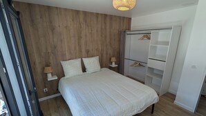 A bedroom with a wooden wall, a bed with a checkered bedspread, bedside lamps, a wardrobe with hanging space, and a ceiling light fixture. - New 3-bedroom villa + SPA (Labenne)