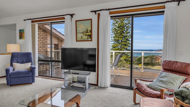 Ein Wohnzimmer mit Fernseher, einem blauen Sessel, einem braunen Ledersofa und einem Glastisch. Es gibt einen Balkon mit Blick auf das Meer und einen Baum.
