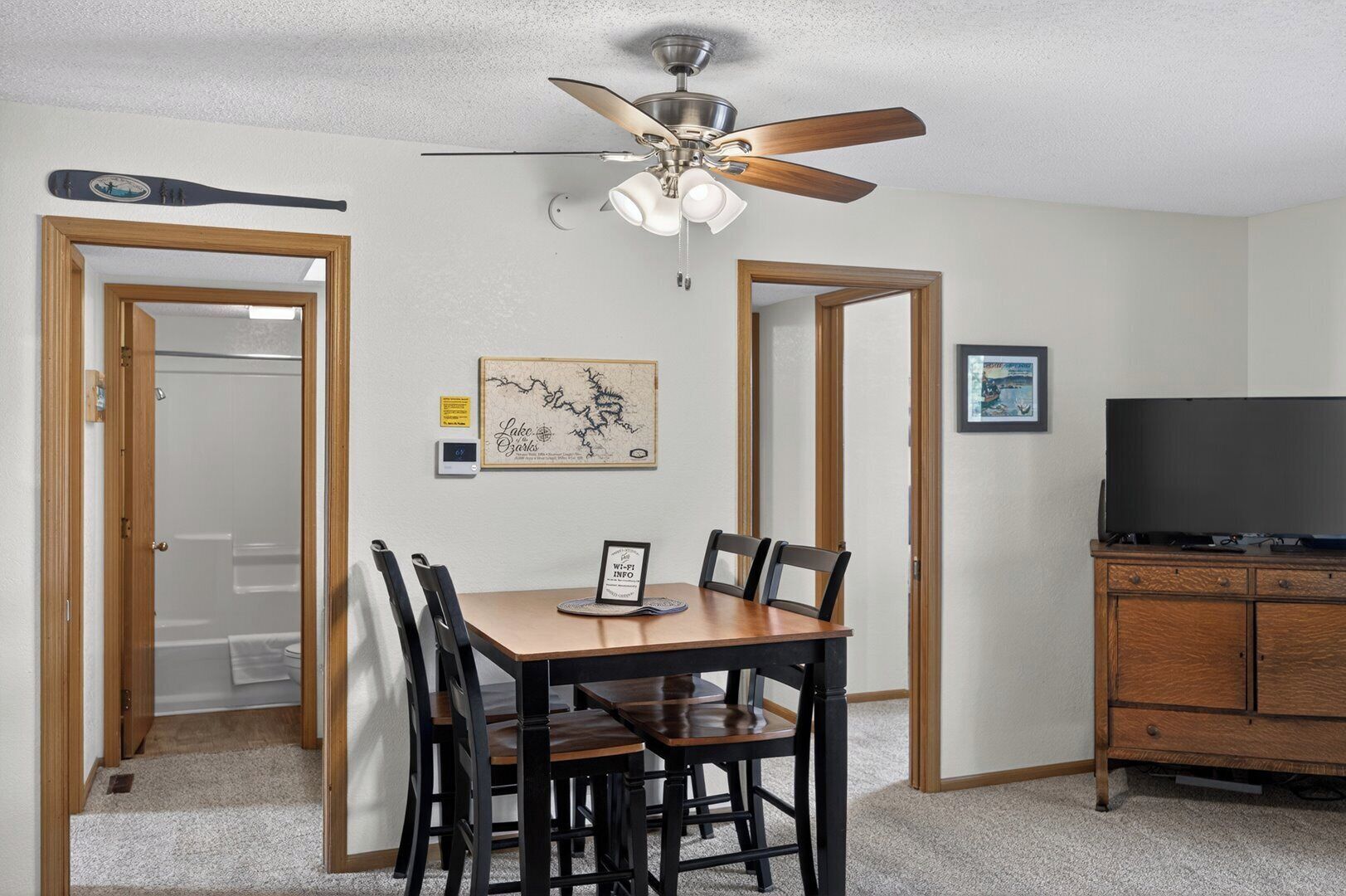 A dining area with a table, chairs, a ceiling fan, a framed artwork, a television, and a cabinet.