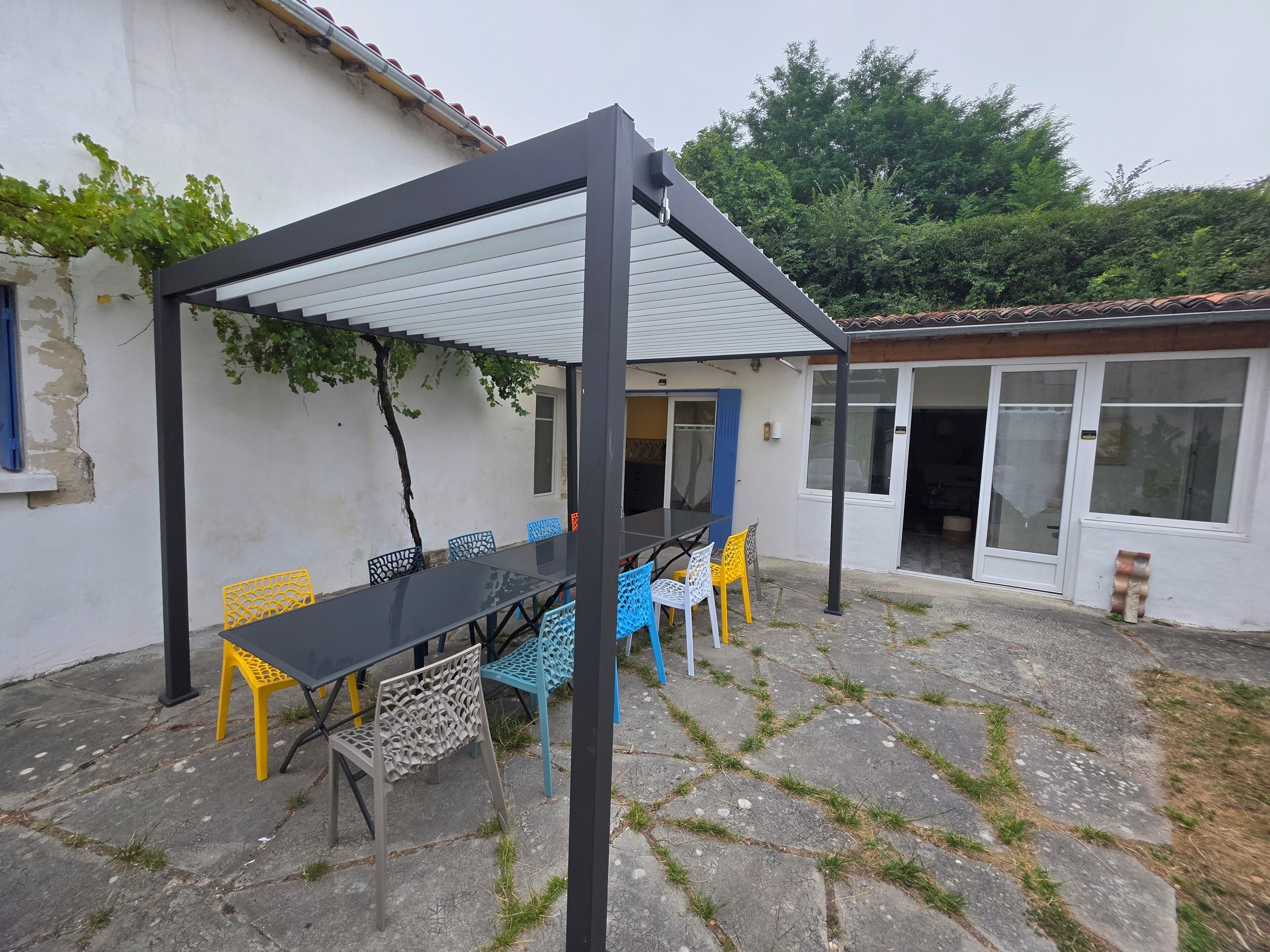 Un patio couvert avec une table et des chaises, une pergola et une vue sur une maison dotée d’une porte-fenêtre coulissante.