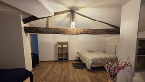 Une chambre avec un plafond en pente, des poutres en bois, un lit recouvert d’une couvre-lit à motifs, une petite table de chevet avec une lampe, une bibliothèque et un vase rempli de fleurs.