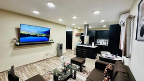 Una sala de estar moderna con un televisor de pantalla plana, una mesa de centro y un sofá. Se observa un área de cocina con estufa y alacenas.