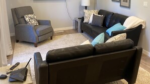 Een woonkamer met een grijze fauteuil, een zwarte bank, een flatscreen televisie aan de muur, een klein bijzettafeltje met een lamp, en een afstandsbediening op een salontafel.