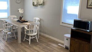 Een eethoek met een houten tafel en witte stoelen, een klok aan de muur en een raam met zonwering.