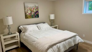 Une chambre à coucher comprenant un lit, deux tables de chevet, une commode, une lampe fixée au mur et une fenêtre donnant sur un paysage verdoyant.