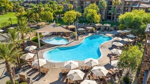 Ein Resort mit einem großen, gebogenen Swimmingpool, umgeben von Liegestühlen und Sonnenschirmen, mit Blick auf die umliegenden Gebäude und die Grünanlage.