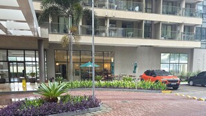 A multi-story building with a paved entrance area, outdoor seating, and a parked car. - Rio Centro/Barra Olímpica Suite (Jacarepaguá Rio de Janeiro)