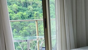 A hotel room with a bed, a balcony with a glass railing, and a view of a forested area. - Rio Centro/Barra Olímpica Suite (Jacarepaguá Rio de Janeiro)
