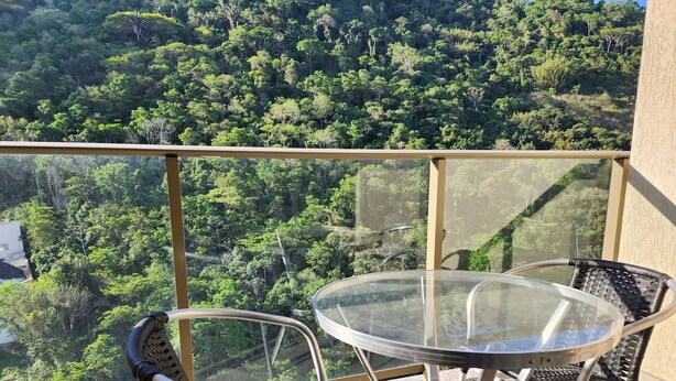 A balcony with a glass table and chairs, overlooking a dense forested hillside. - Rio Centro/Barra Olímpica Suite (Jacarepaguá Rio de Janeiro)