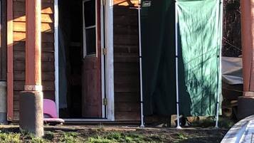 A wooden cabin with a sliding glass door, a green tarp covering part of the structure, and a pink chair outside.