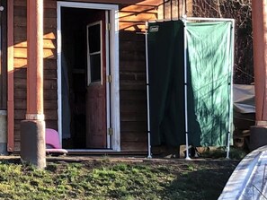 Une cabane en bois avec une porte coulissante en verre, une bâche verte recouvrant une partie de la structure, et une chaise rose à l’extérieur.