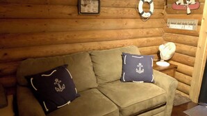 Interior acolhedor de uma cabana com parede de toras de madeira, um sofá estofado com almofadas temáticas náuticas, uma mesinha com um ventilador e uma placa de boas-vindas com ilustração de um guaxinim.