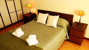 Una cama bien hecha con almohadas y toallas dobladas, dos mesitas de noche con lámparas, y un suelo de madera.