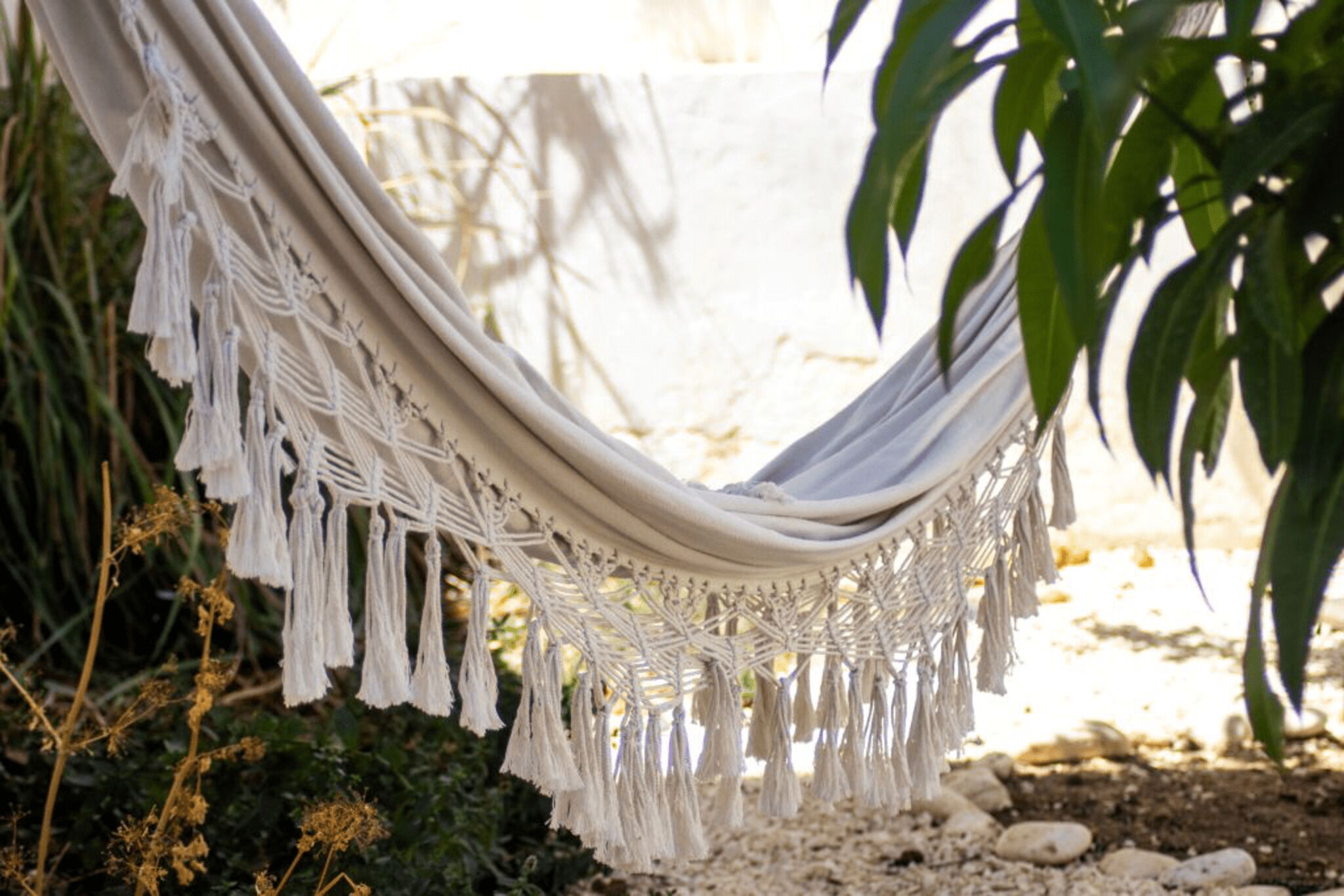 A hammock with macrame fringe, set against a natural backdrop with plants and rocks.