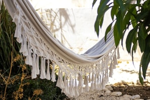A hammock with macrame fringe, set against a natural backdrop with plants and rocks.