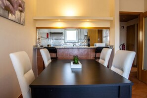 A dining area with a long table, four chairs, a small plant centerpiece, and a kitchen in the background.