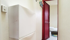 Un couloir avec un réfrigérateur blanc, une porte rouge et un sol carrelé, qui mène à une chambre à coucher où se trouvent un lit et un tapis.