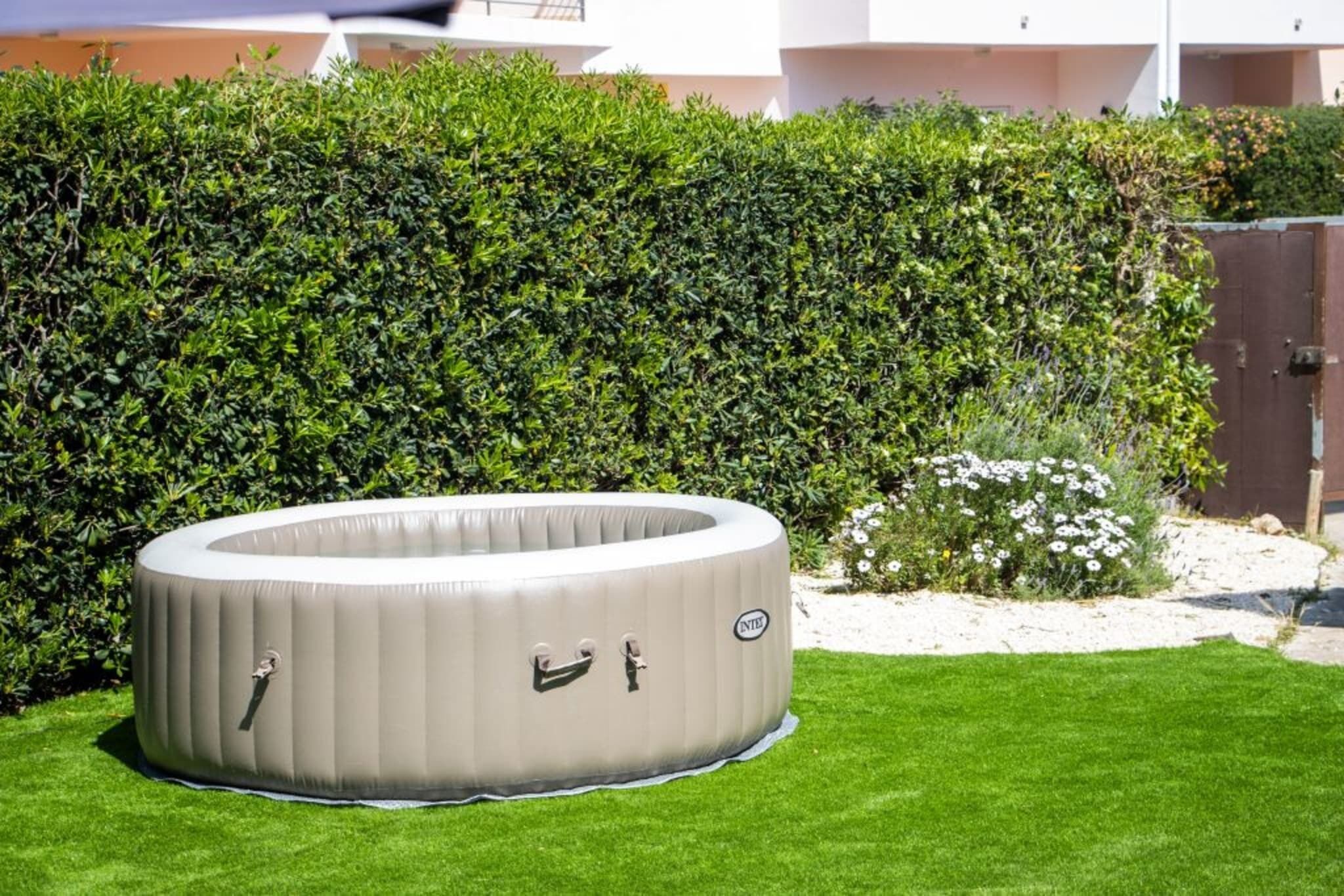 An inflatable pool in a garden with a hedge and a building in the background.