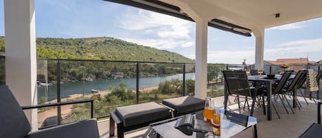 A balcony with a glass table, chairs, and a sofa, overlooking a river and hills.
