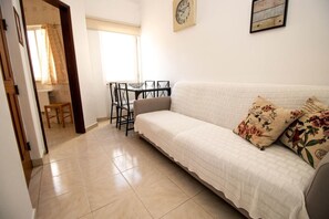 Ein Zimmer mit einem weißen Sofa, einem Esstisch mit Stühlen, einer Wanduhr und einem Fenster mit Vorhängen.