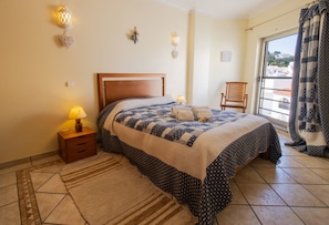 A bedroom with a bed, nightstand, lamp, and a view of the outside.