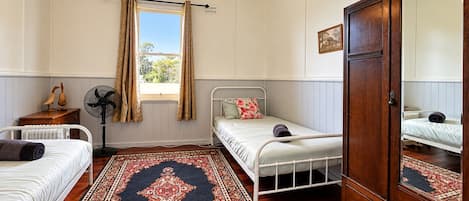 Une chambre avec deux lits simples, une armoire en bois, un radiateur, un ventilateur, une fenêtre avec des rideaux et un tapis à motifs.