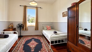 A room with two single beds, a wooden wardrobe, a radiator, a fan, a window with curtains, and a patterned rug.