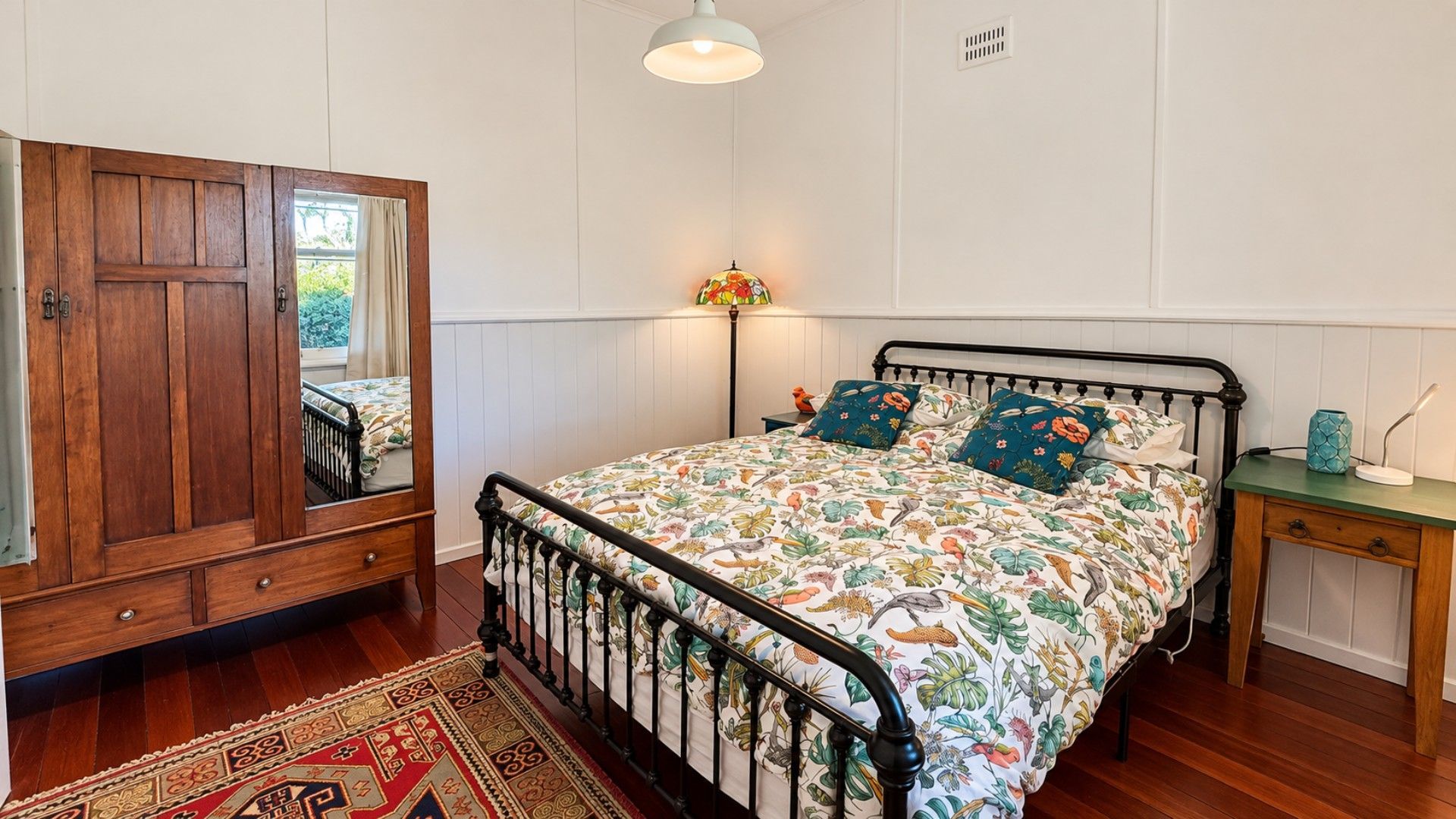 A bedroom with a wooden wardrobe, a bed with floral bedding, a nightstand, and a floor lamp.