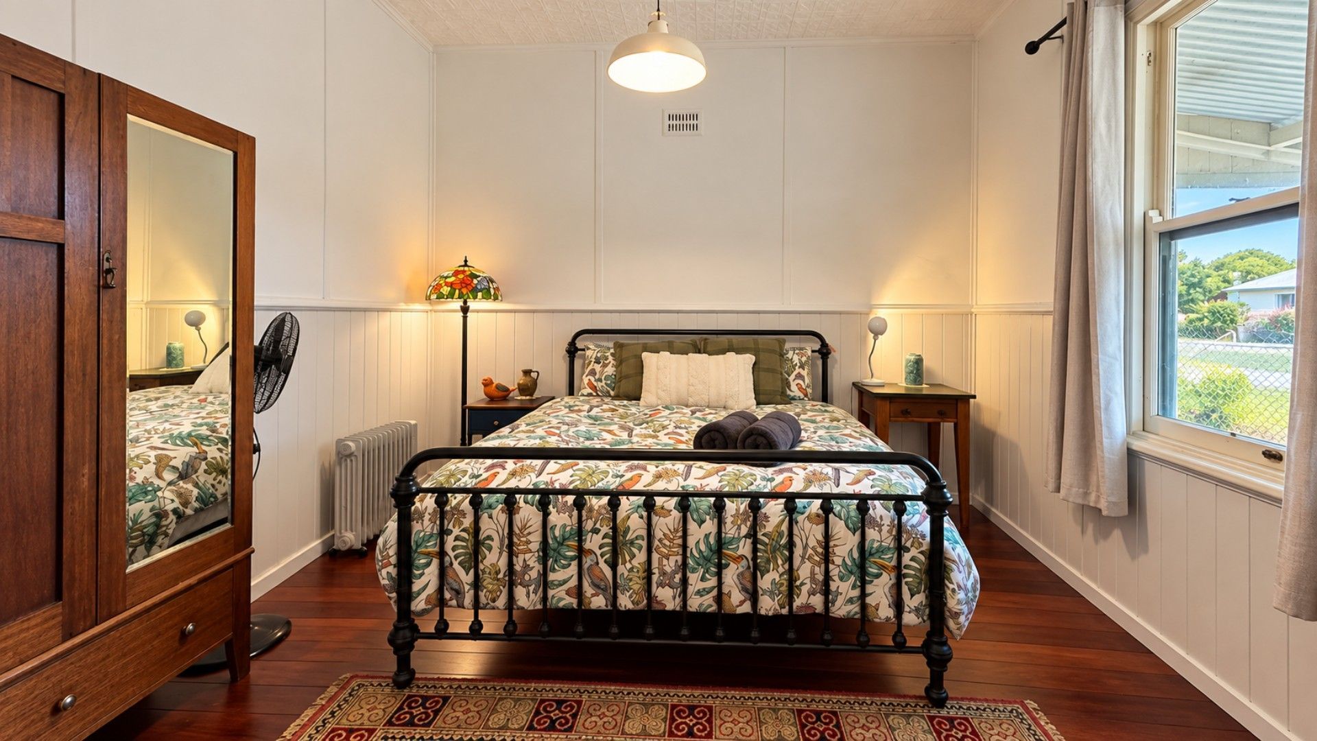 A bedroom with a bed, a nightstand, a mirror, a radiator, a window with curtains, and a fan.
