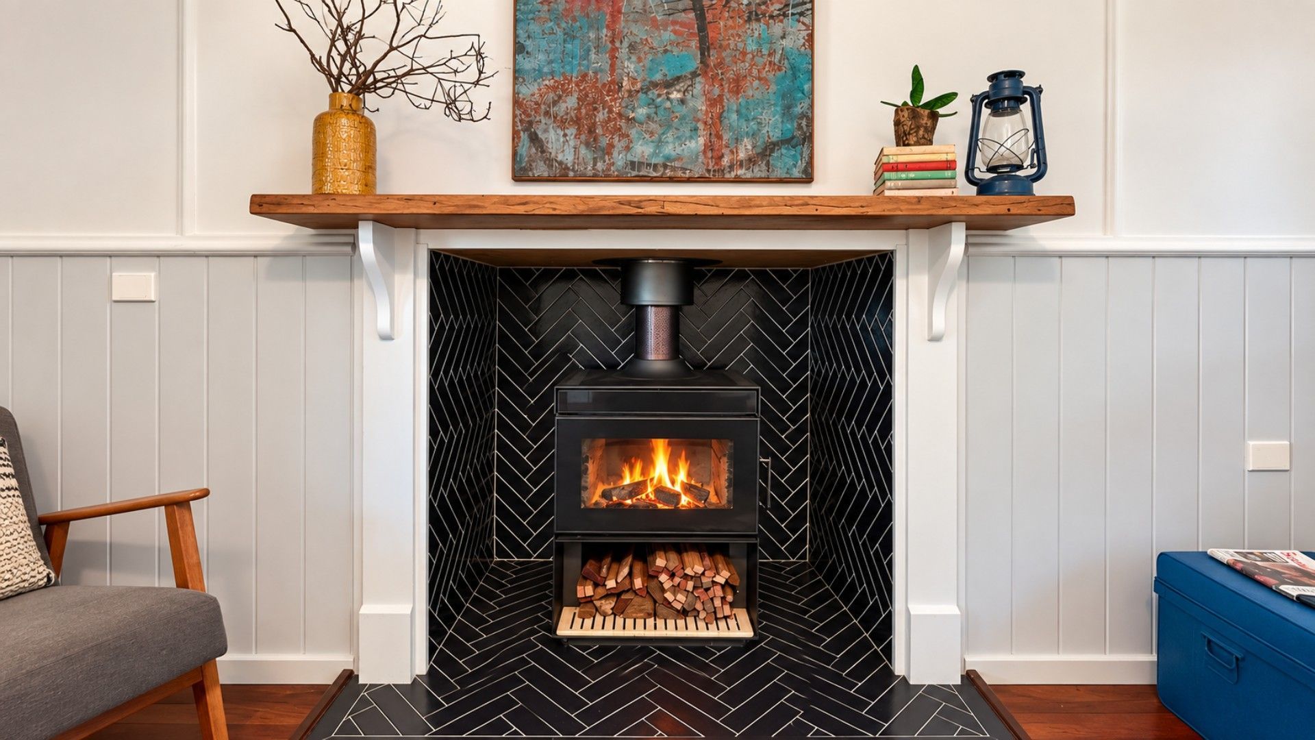 A fireplace with a herringbone-patterned surround, a mantel shelf with decorative items, and a chair to the left.